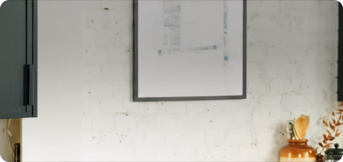 White brick wall with a framed blank picture and a brown jar holding wooden utensils.