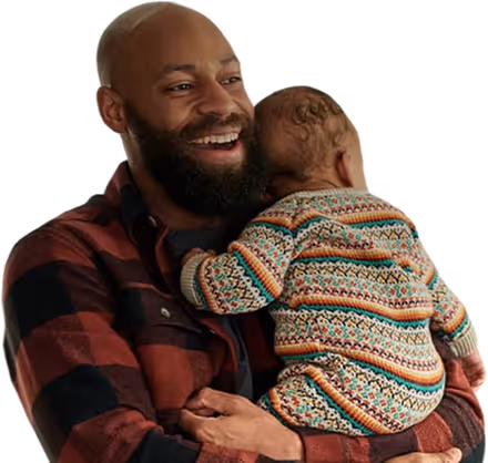 Smiling man in plaid shirt holding a baby wearing a colorful knit sweater.