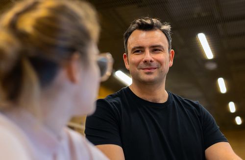 Un homme souriant en t-shirt noir parlant à une femme floue portant des lunettes.