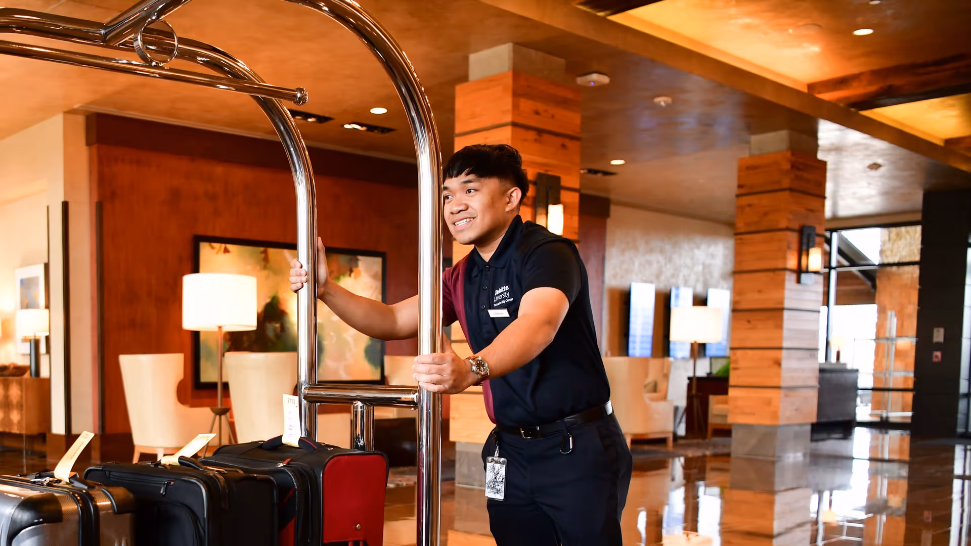 Hotel bellman smiling while pushing a luggage cart with suitcases in a warmly lit lobby.