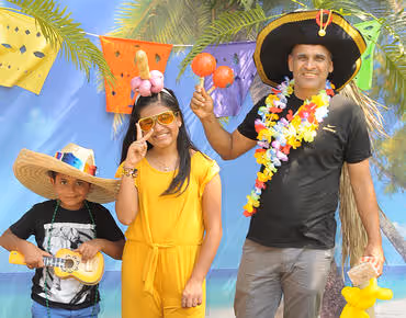 Smiling man wearing a large sombrero and floral lei holding maracas, with a girl in yellow outfit and sunglasses, and a boy in a straw hat playing a small guitar, standing in front of colorful decorations and palm leaves.