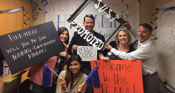 Five smiling coworkers celebrating a promotion with signs reading 'Johnothan will you be our training & communication manager?', 'Yes', and 'Welcome to the team!' in an office decorated for the occasion.