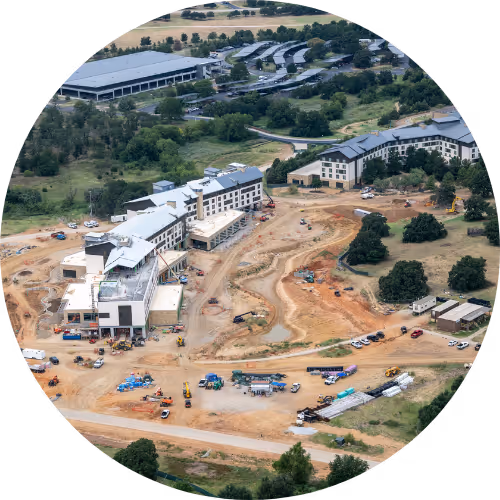 Aerial view of a large construction site with partially completed multi-story buildings, construction vehicles, and dirt roads surrounded by green trees and an adjacent parking area.