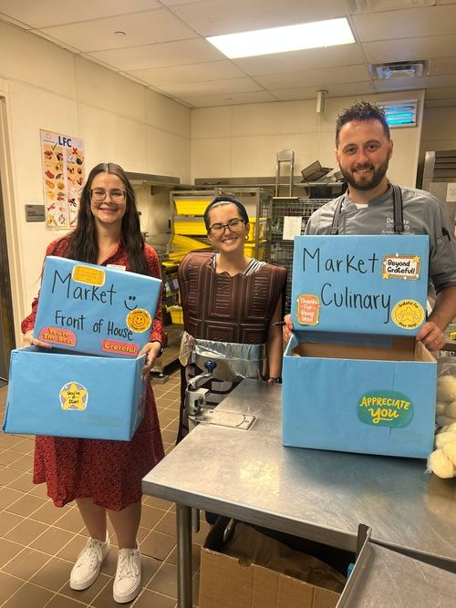 Three smiling people in a kitchen, one wearing a chocolate bar costume, holding blue boxes labeled Market Front of House and Market Culinary with positive stickers.