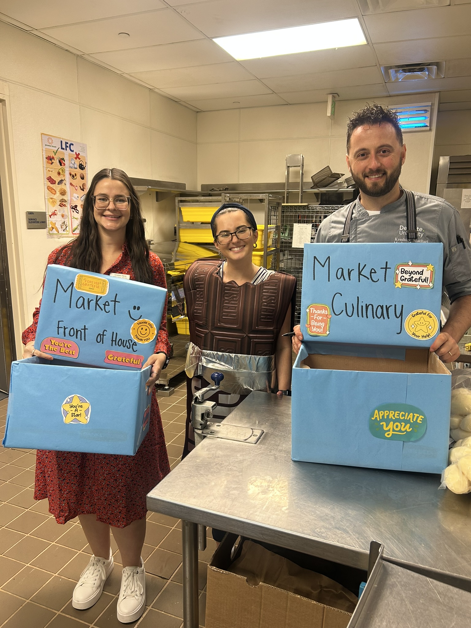 Three smiling people in a kitchen, one wearing a chocolate bar costume, holding blue boxes labeled Market Front of House and Market Culinary with positive stickers.
