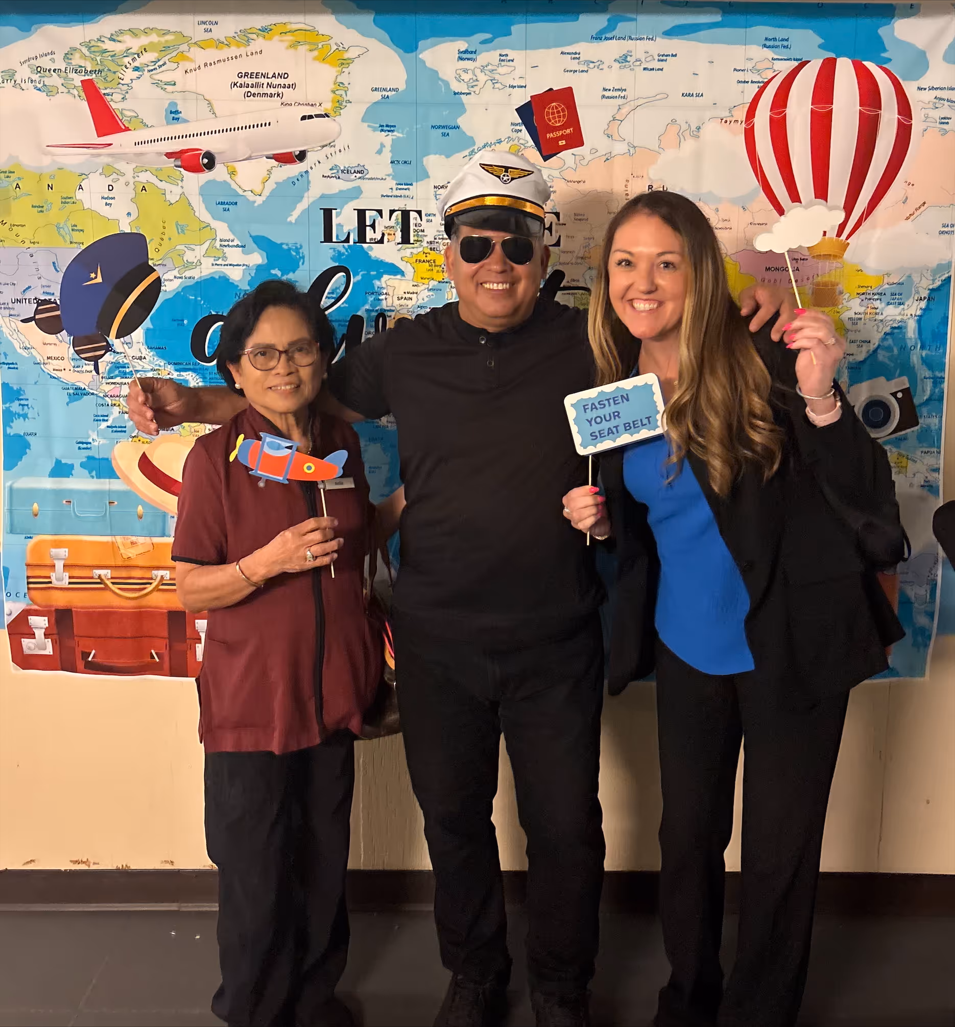 Three people smiling in front of a world map backdrop with travel-themed props including a pilot hat, toy airplane, 'Fasten Your Seat Belt' sign, and a hot air balloon cutout.