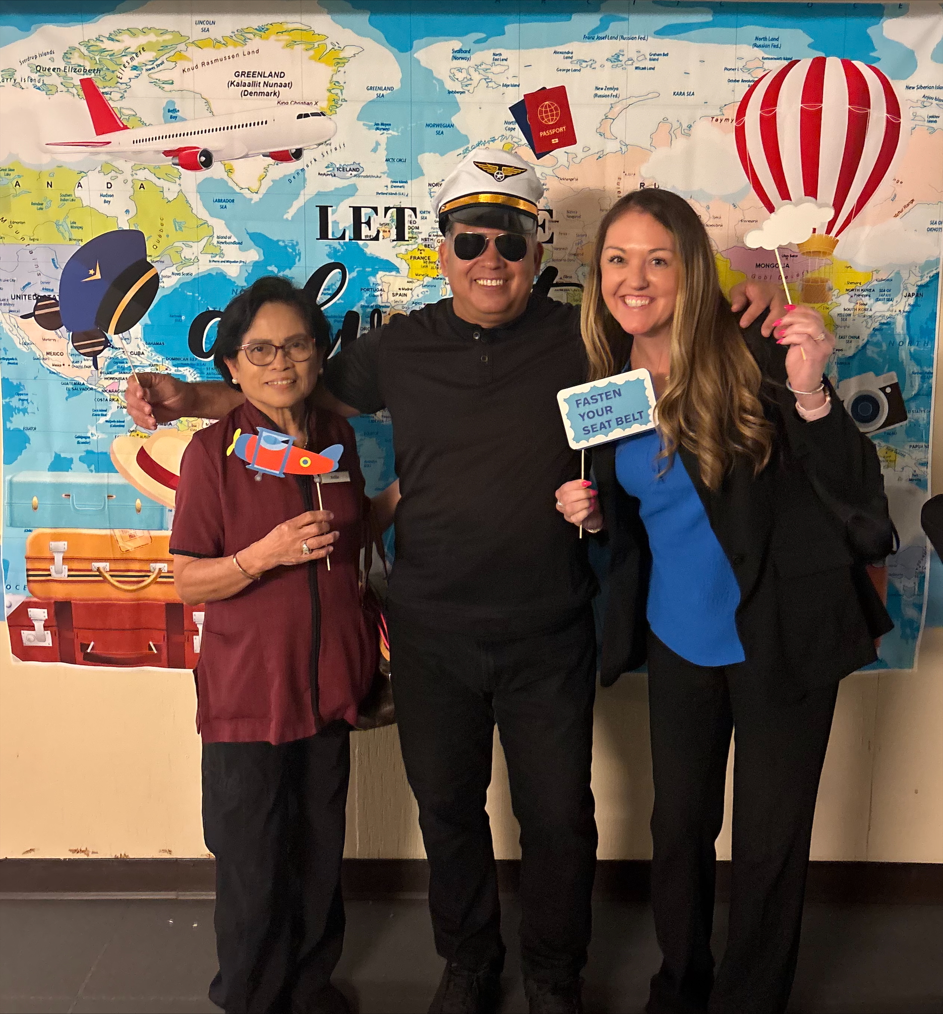 Three people smiling in front of a world map backdrop with travel-themed props including a pilot hat, toy airplane, 'Fasten Your Seat Belt' sign, and a hot air balloon cutout.