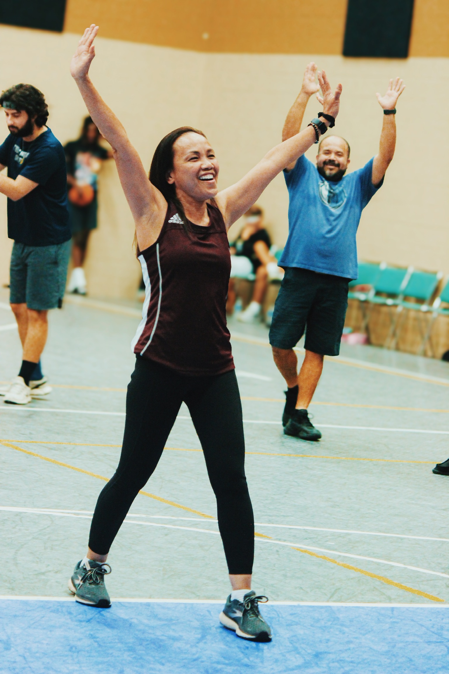 A woman in athletic wear with arms raised smiles during an indoor group exercise class.