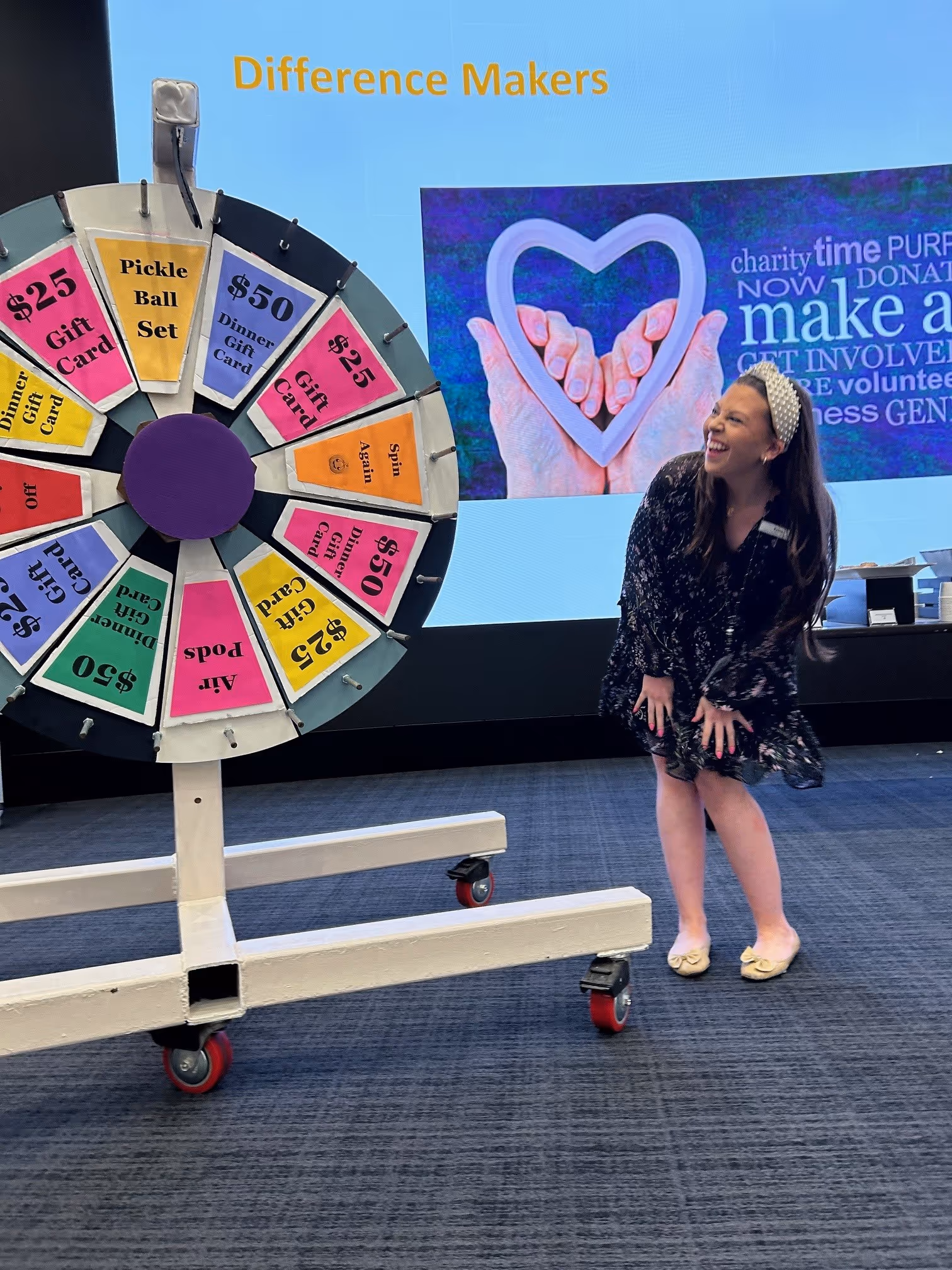 A woman laughing next to a colorful prize wheel with sections offering gift cards and a pickle ball set in a room with a 'Difference Makers' sign and charity-themed presentation on the screen.