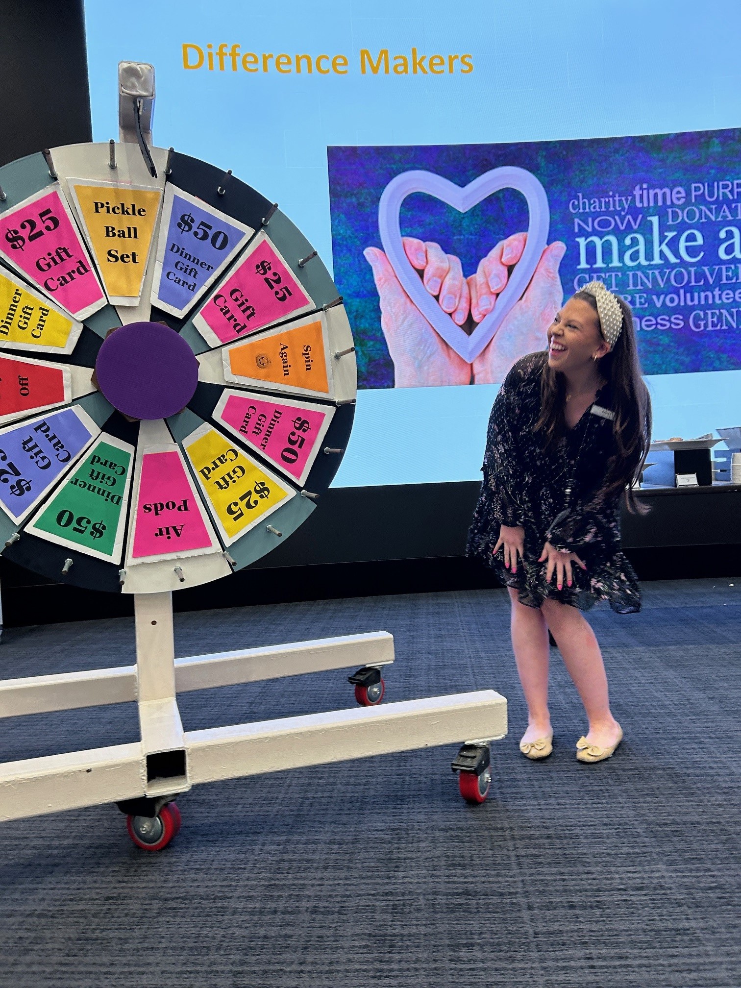 A woman laughing next to a colorful prize wheel with sections offering gift cards and a pickle ball set in a room with a 'Difference Makers' sign and charity-themed presentation on the screen.