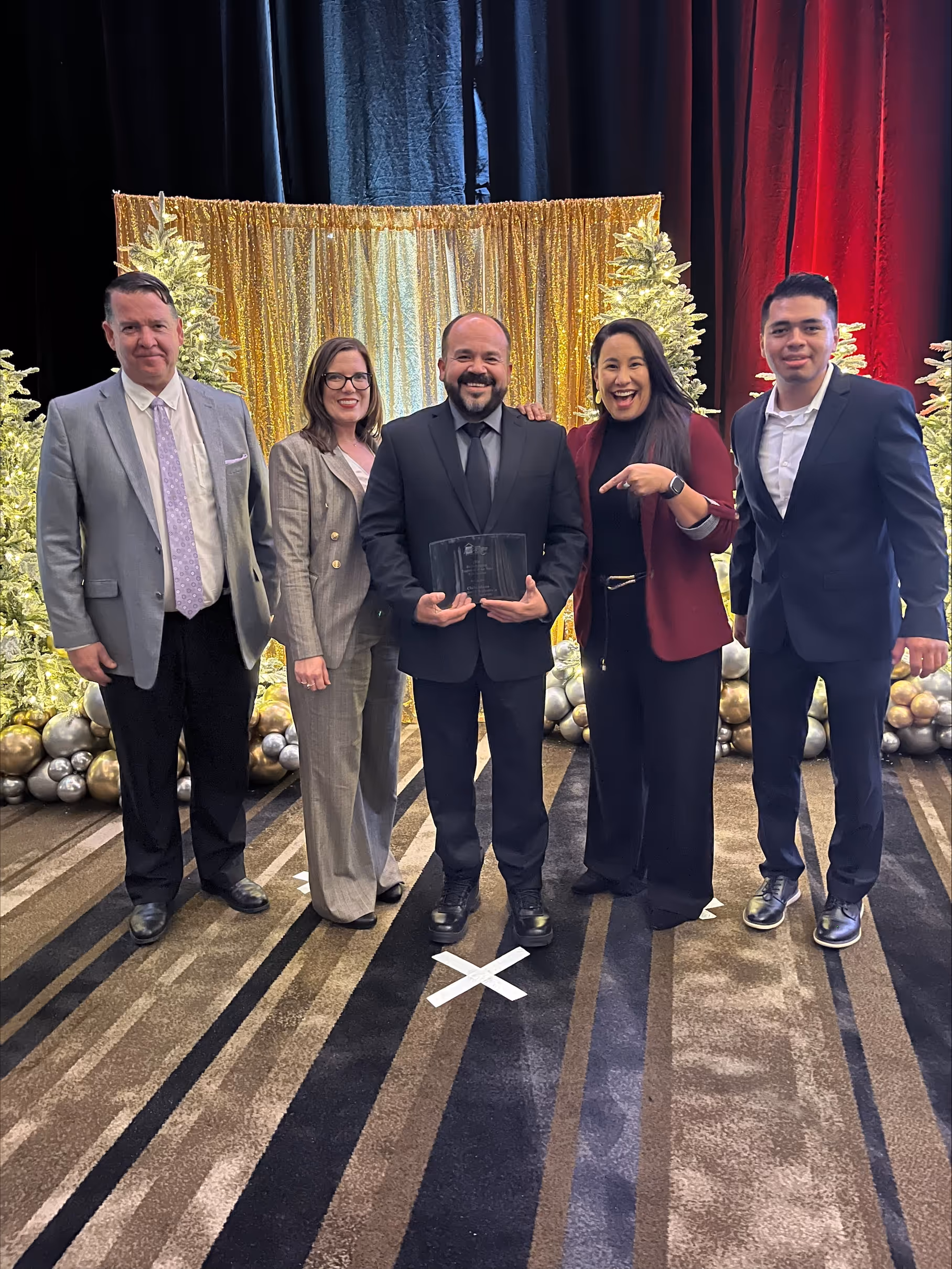 Group of five people in formal attire standing on a carpet with gold and silver holiday decorations and a gold sequin backdrop, one man in the center holding an award plaque.