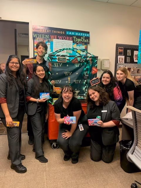 Six diverse women smiling and holding candy bars in an office decorated with a green and black themed cart and a motivational sign on the wall.