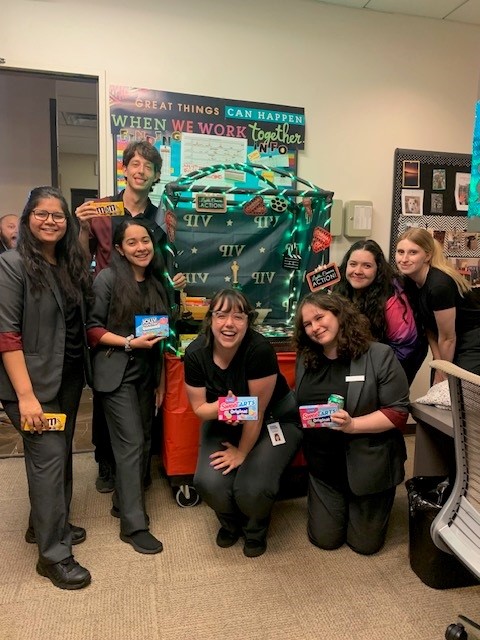 Six diverse women smiling and holding candy bars in an office decorated with a green and black themed cart and a motivational sign on the wall.