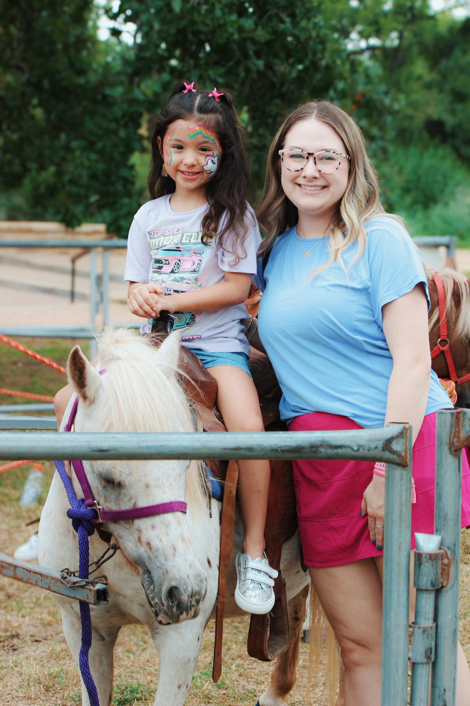 Smiling young girl with unicorn face paint riding a white pony beside a woman in glasses and a blue shirt.