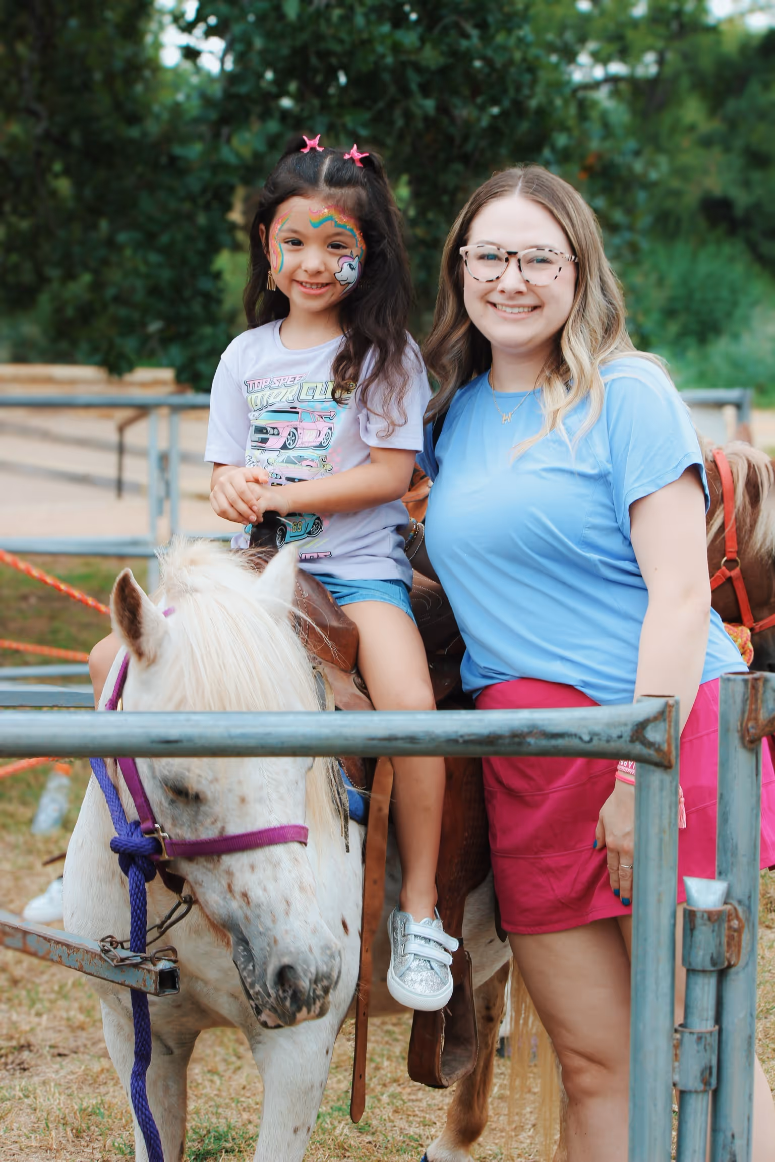 Smiling young girl with unicorn face paint riding a white pony beside a woman in glasses and a blue shirt.