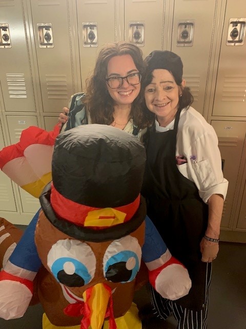 Two smiling women standing in front of lockers with an inflatable turkey wearing a black top hat and red bow tie in the foreground.