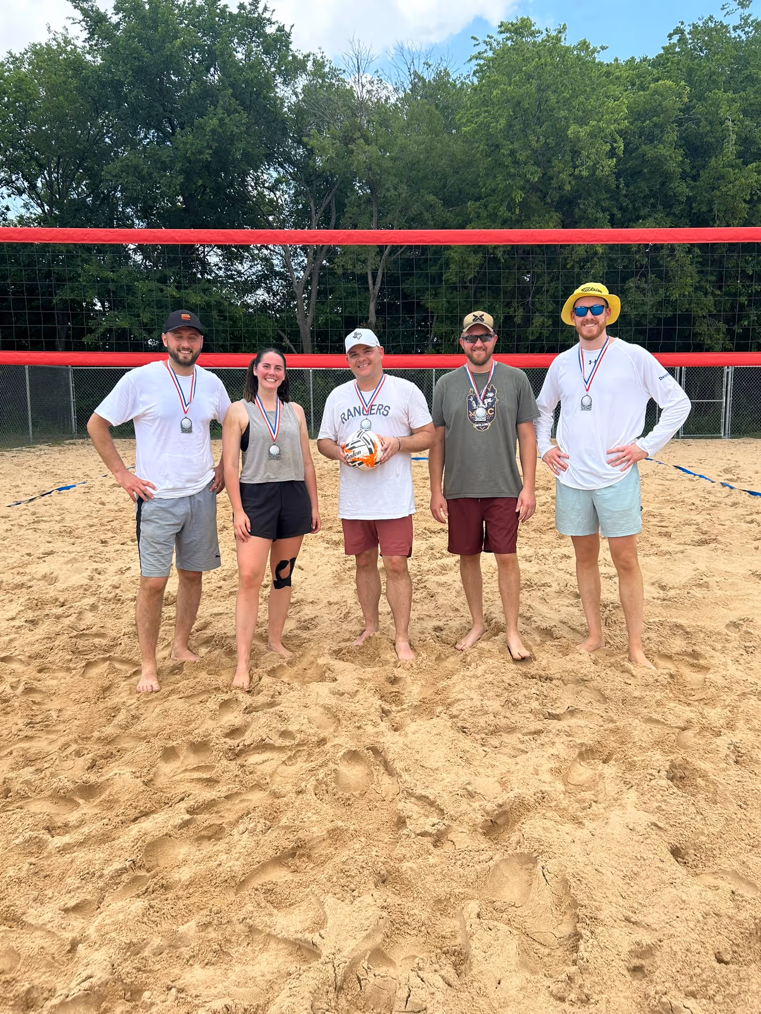 Group of five smiling people standing barefoot on sand volleyball court wearing medals, with a volleyball held by the middle person.