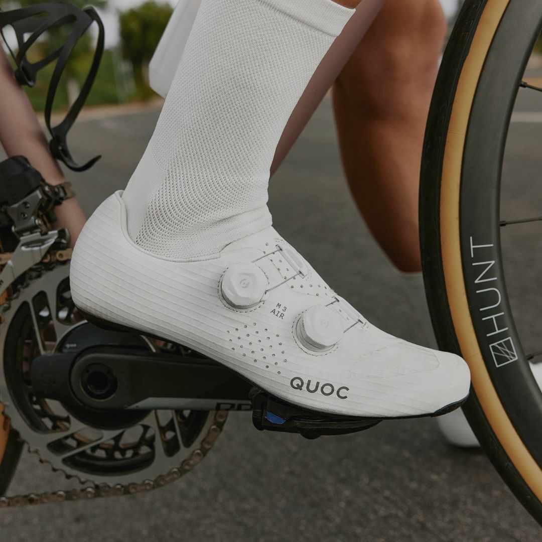 White cycling shoe clipped into bike pedal with visible drivetrain and carbon wheel