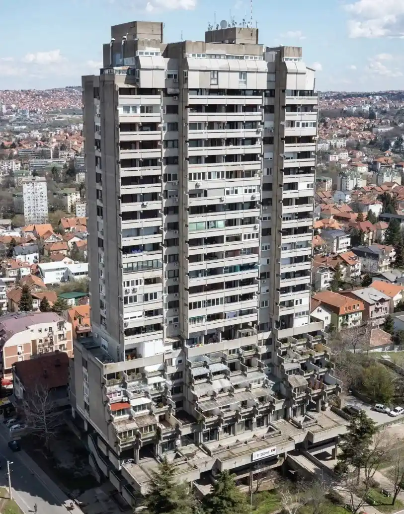 Visoka, siva betonska stambena zgrada sa više balkona u gusto naseljenoj stambenoj zoni pod delimično oblačnim nebom.