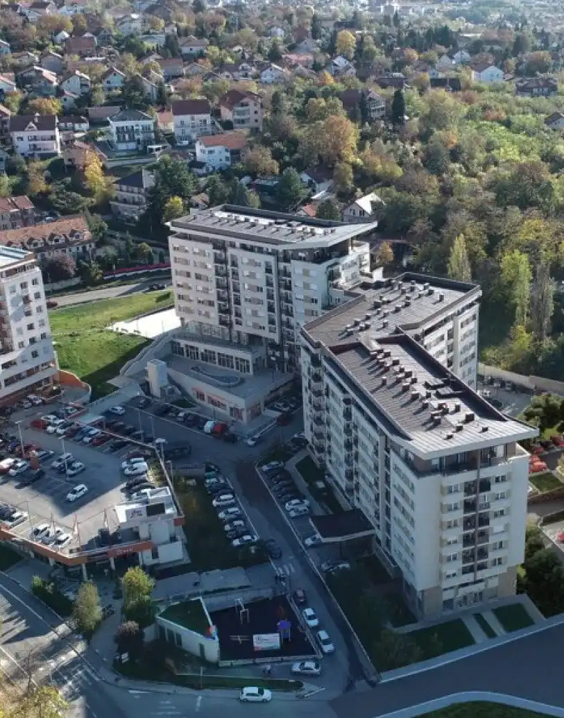 Pogled iz vazduha na stambeni kompleks sa dve velike višespratne stambene zgrade okružene parkiranim automobilima i zelenilom.
