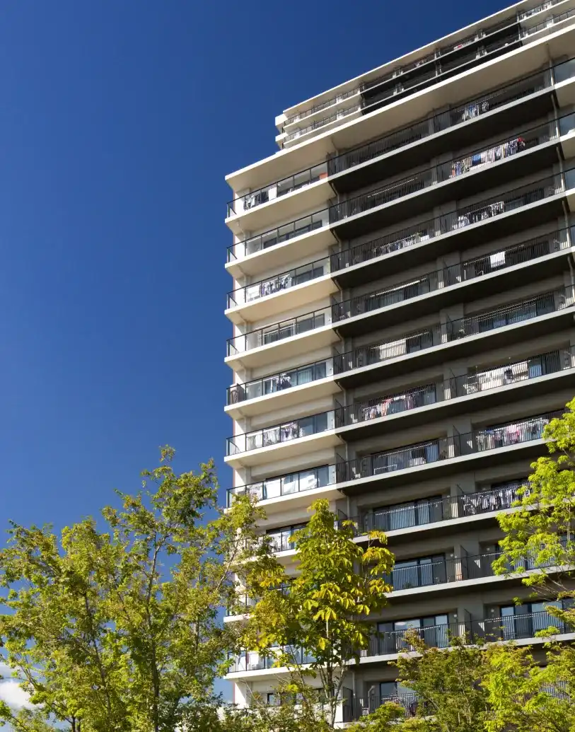 Visoka stambena zgrada sa balkonima uz vedro plavo nebo i zeleno drveće u prvom planu.