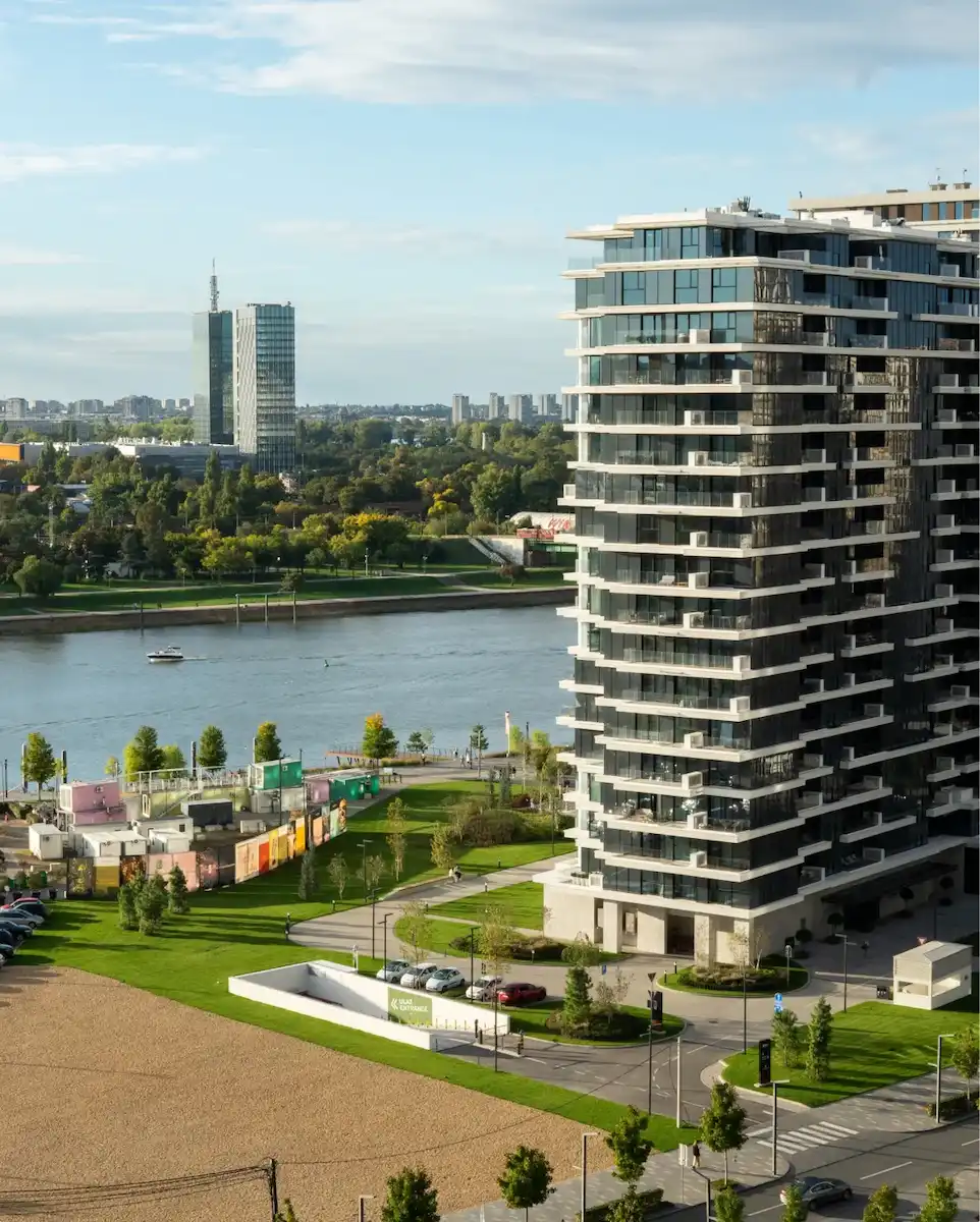 Moderna stambena zgrada pored reke sa staklenim balkonima sa pogledom na reku i zeleni park pod delimično oblačnim nebom.
