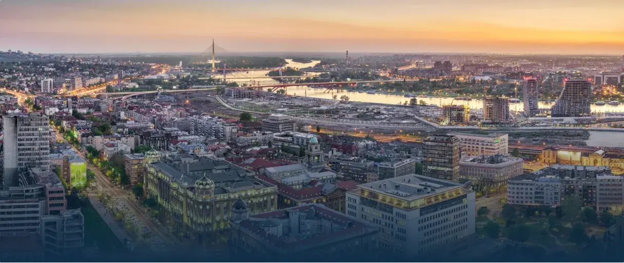 Panoramski gradski pejzaž u zalazak sunca koji prikazuje nebodere, mostove preko reke i osvetljene ulice.