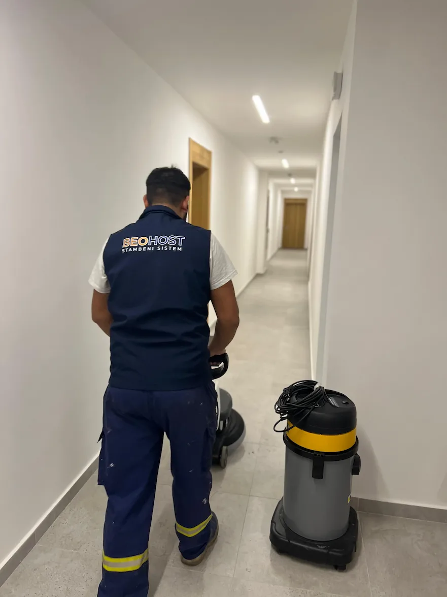 Osoba u uniformi BEOHOST-a upravlja mašinom za čišćenje poda u belom hodniku sa zatvorenim drvenim vratima.