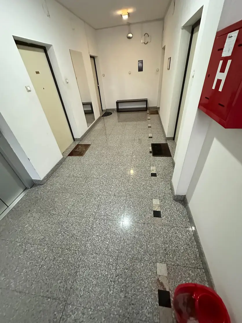 Unutrašnji hodnik sa bež vratima, ogledalom na zidu, crvenom kutijom za vatrogasno crevo i malom klupom na kraju.