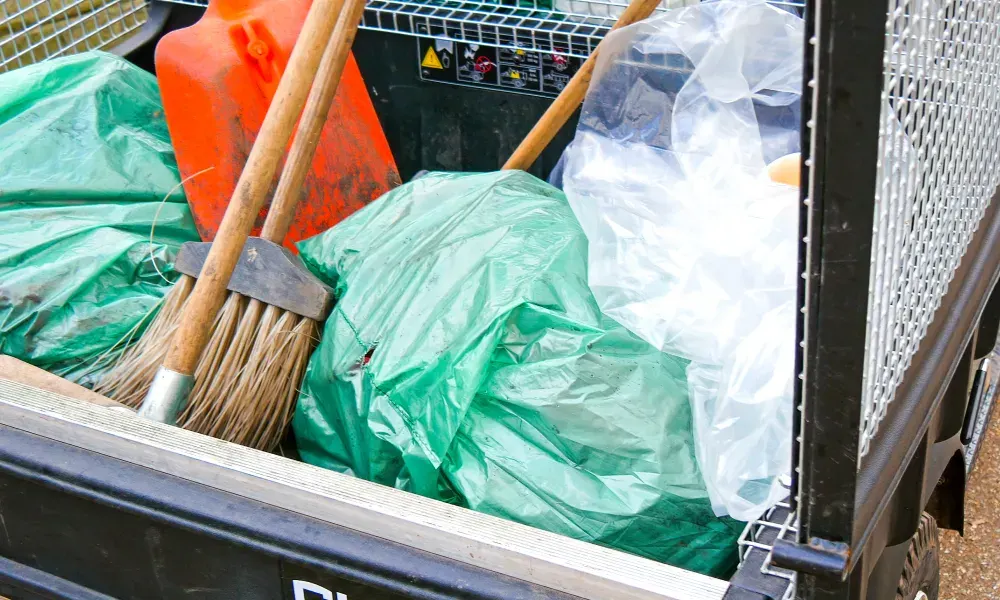 Zadnji deo komunalnog vozila natovaren zelenim kesama za smeće, metlama, narandžastim bojlerom za gas i belom plastičnom kesom. Zadnji deo komunalnog vozila natovaren zelenim kesama za smeće, metlama, narandžastim bojlerom za gas i belom plastičnom kesom.