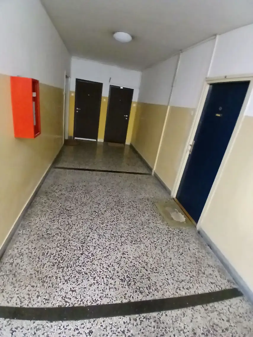 Unutrašnji hodnik sa teraco podom, bež i belim zidovima, troja zatvorena vrata i crvenom kutijom za protivpožarni aparat na levom zidu.