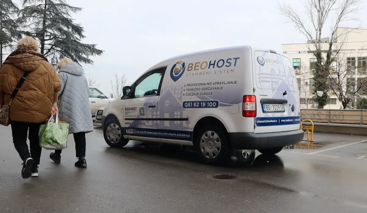 Beli kombi kompanije Beohost parkiran na mokroj ulici, a u blizini su hodale dve starije žene noseći torbe.