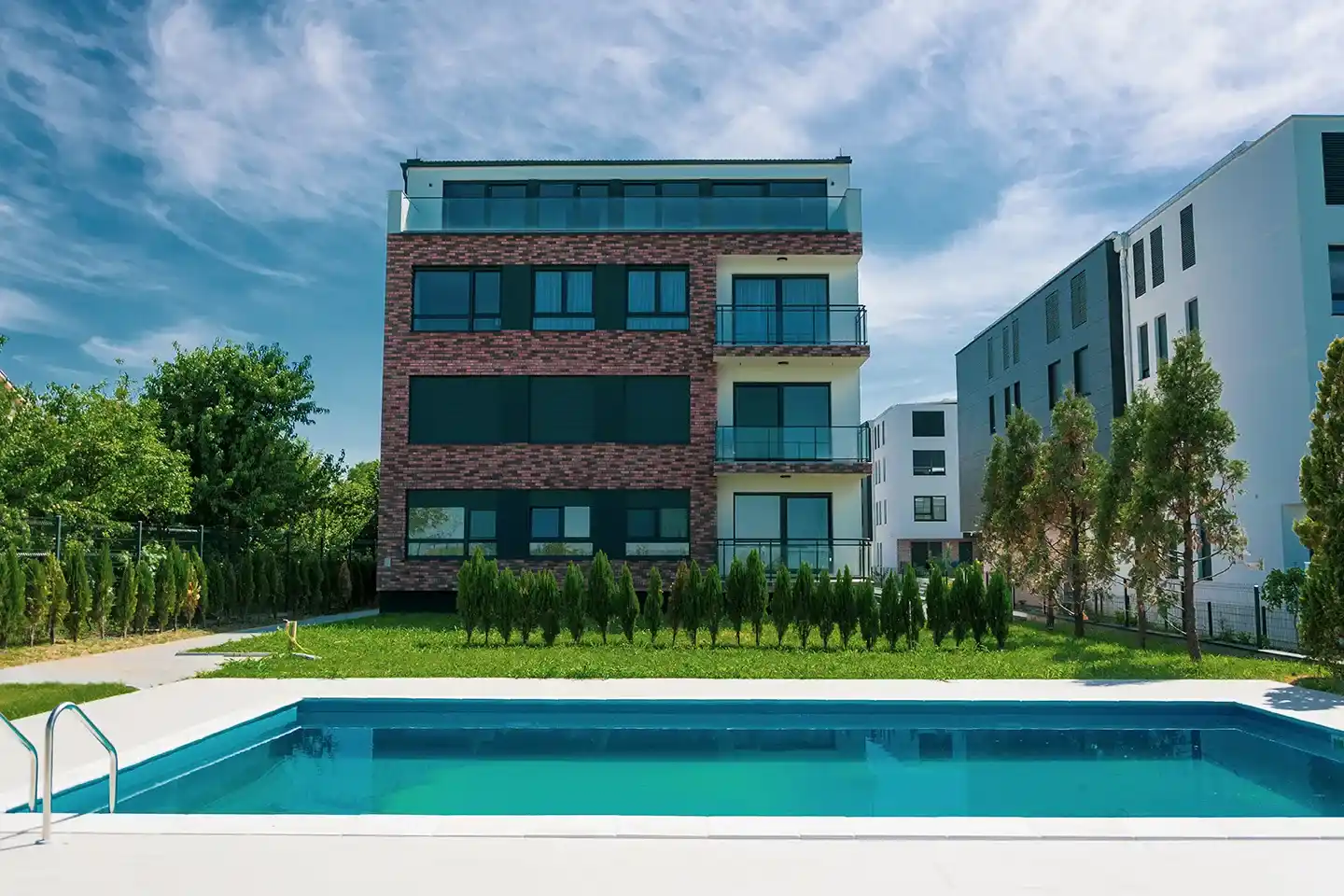 Moderna trospratna stambena zgrada sa balkonima i velikim prozorima, zelenim travnjakom i pravougaonim bazenom u prvom planu, pod delimično oblačnim nebom.