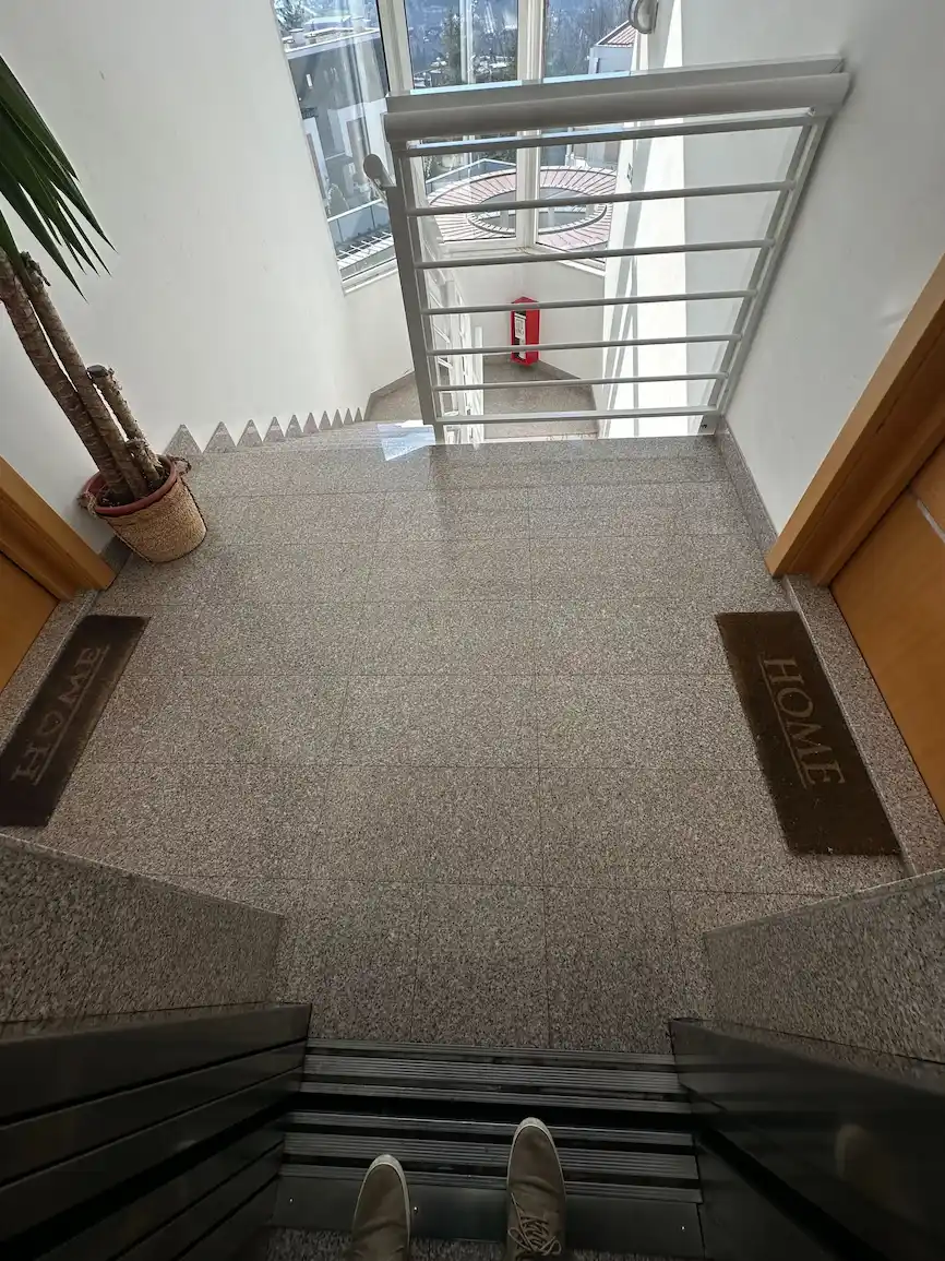 Pogled kroz otvorena vrata koji prikazuje popločan podest sa dva otirača sa natpisima „LOVE“ i „HOME“, pored saksije sa biljkom, sa pogledom na stepenište sa metalnom zaštitnom kapijom i protivpožarnim aparatom na dnu.