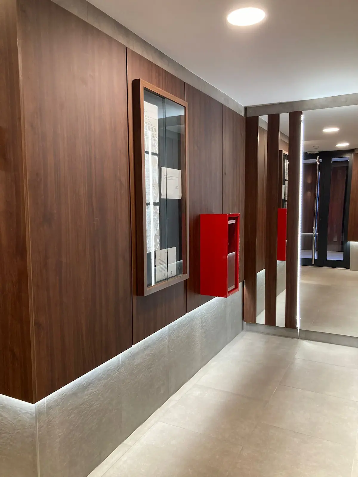 Moderni hodnik sa zidovima obloženim drvenim panelima, crvenom kutijom za zaštitu od požara, staklenom vitrinom i ogledalom od poda do plafona koje odražava prostor.