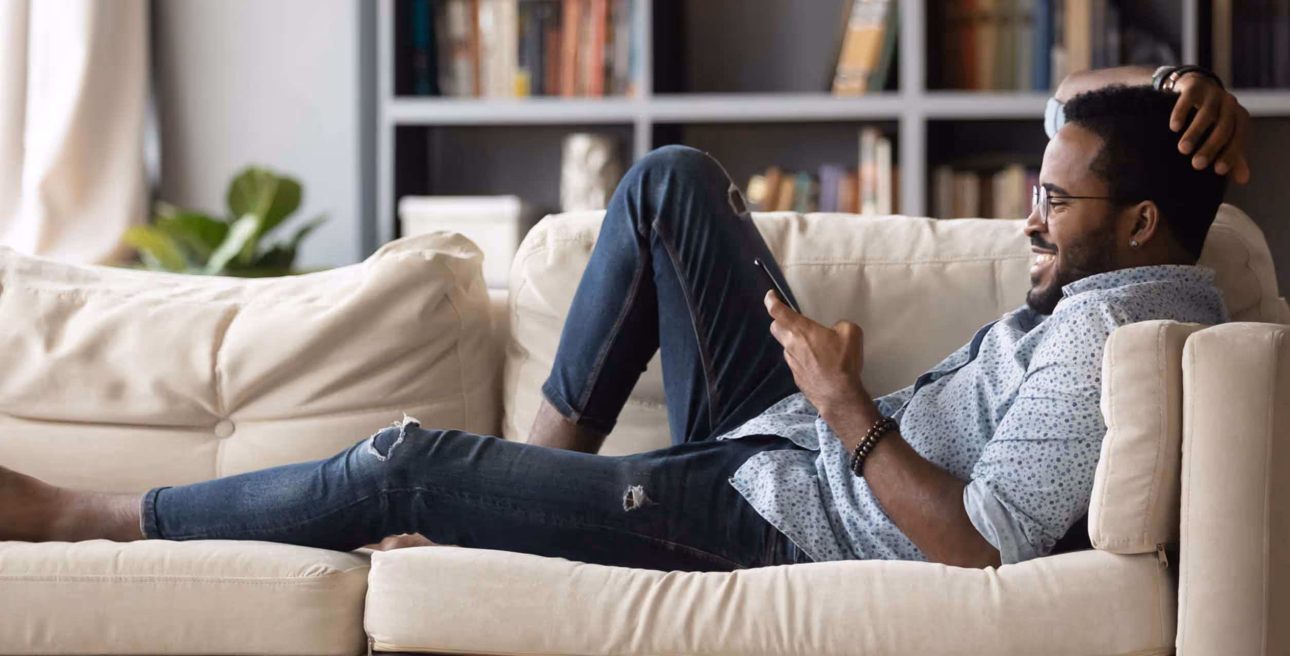 Smiling man in glasses and patterned shirt reclining on beige couch looking at his smartphone.