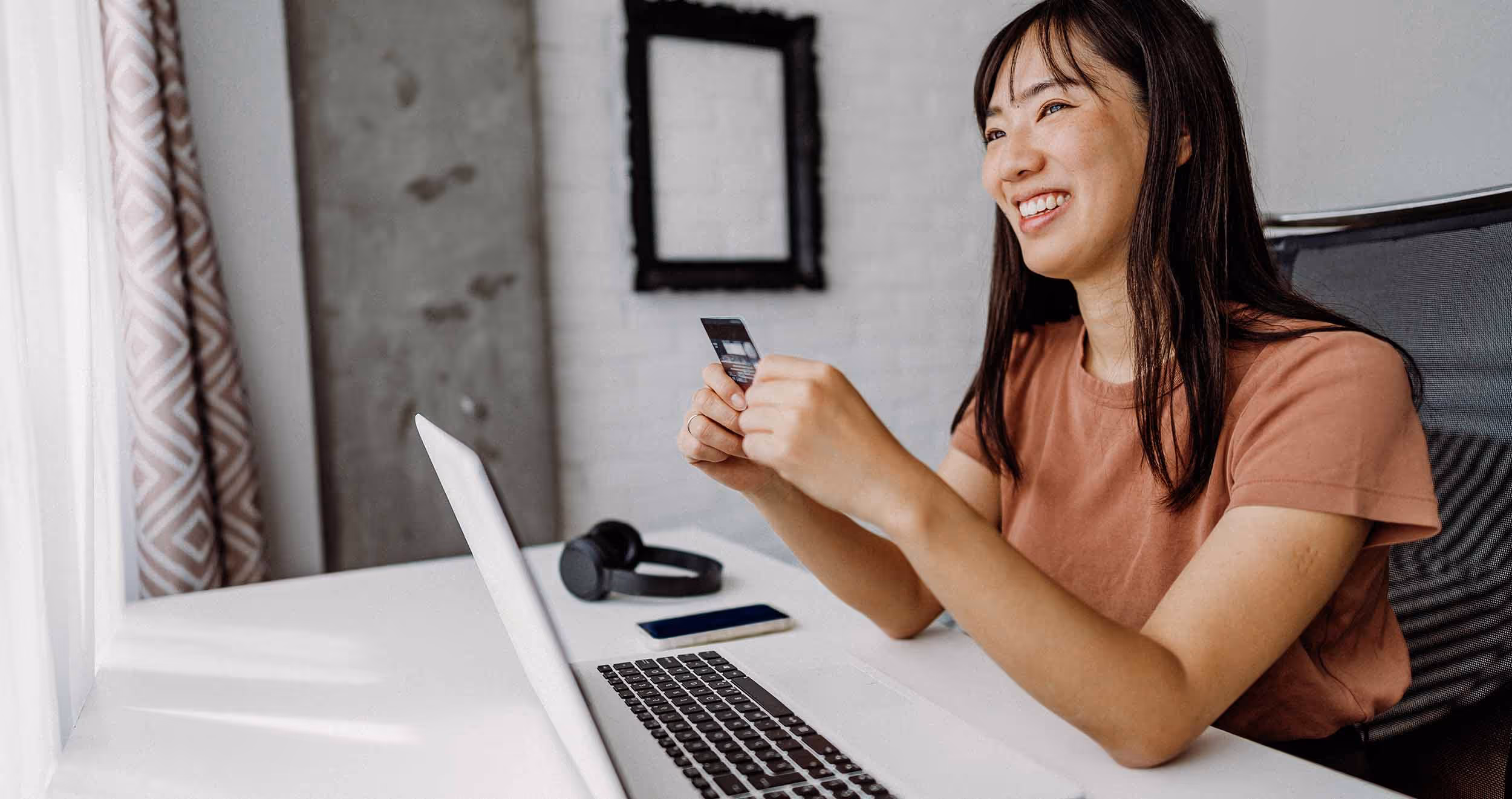 Smiling woman sitting at a desk holding a credit card with an open laptop, phone, and headphones nearby.