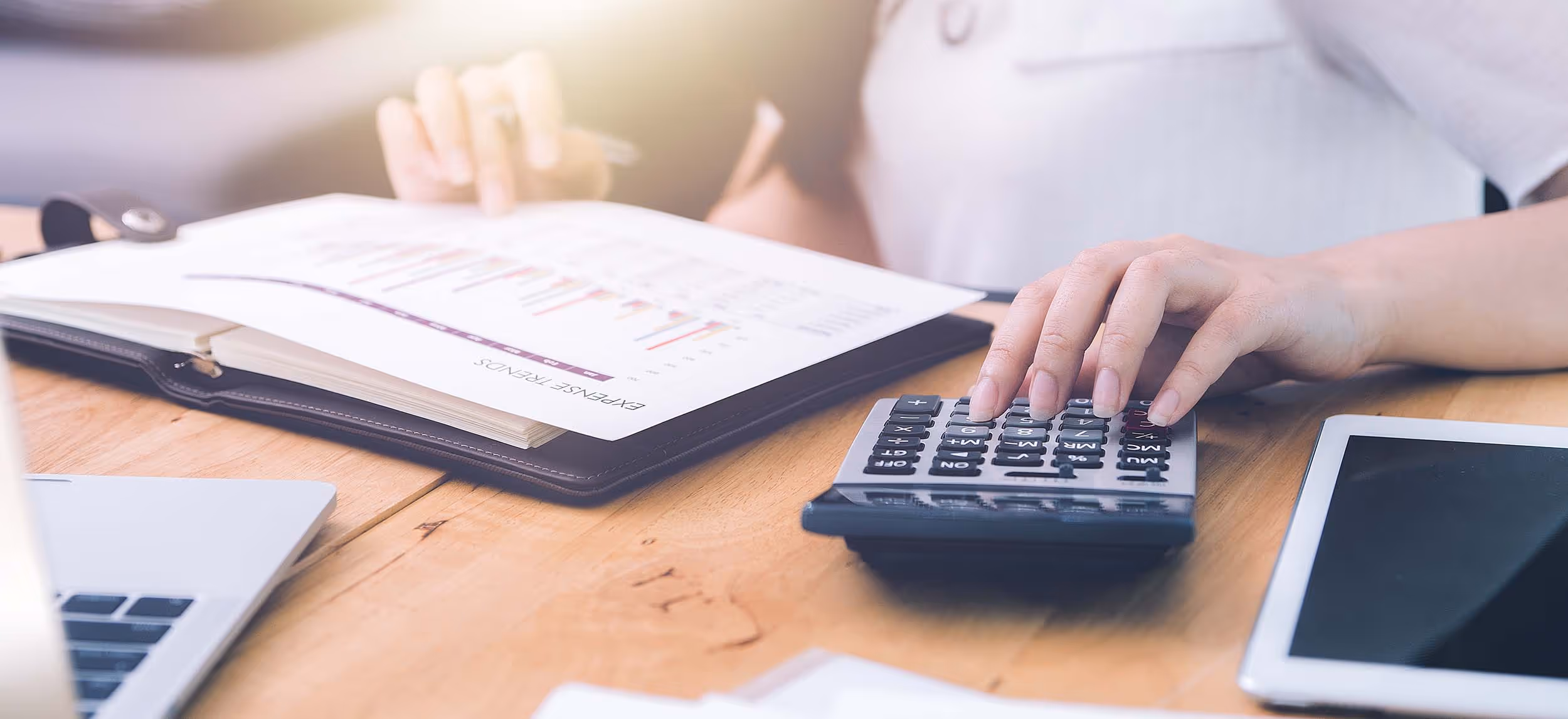 Person using a calculator and reviewing a report with expense trends at a wooden desk with a laptop and tablet nearby.