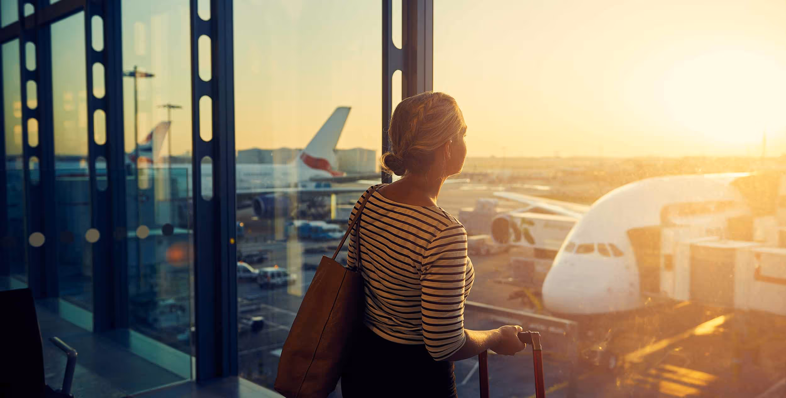 Woman with striped shirt and tote bag holding suitcase handle looking out airport window at airplanes during sunset.