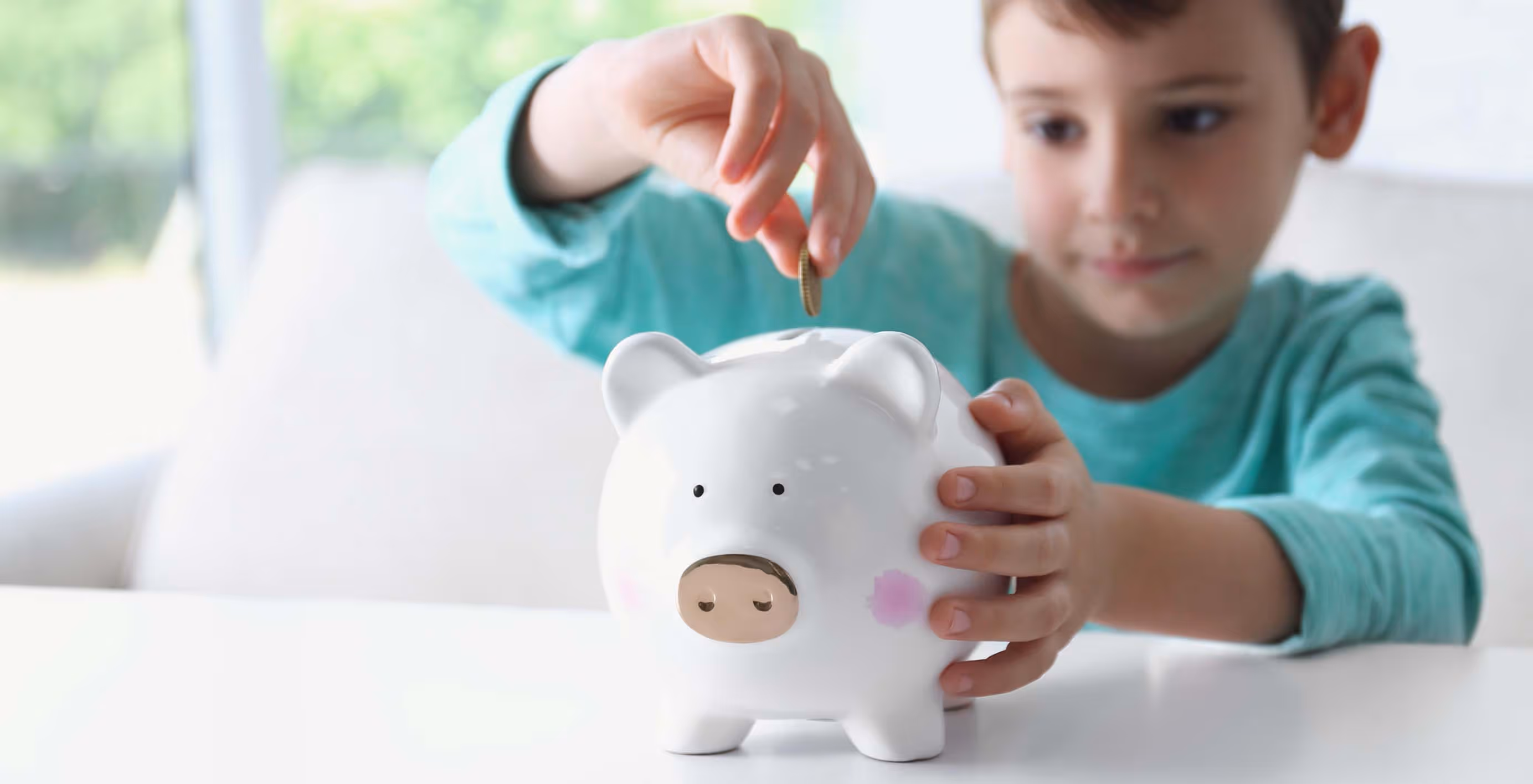 Child in turquoise shirt placing a coin into a white piggy bank with pink cheeks on a white table.