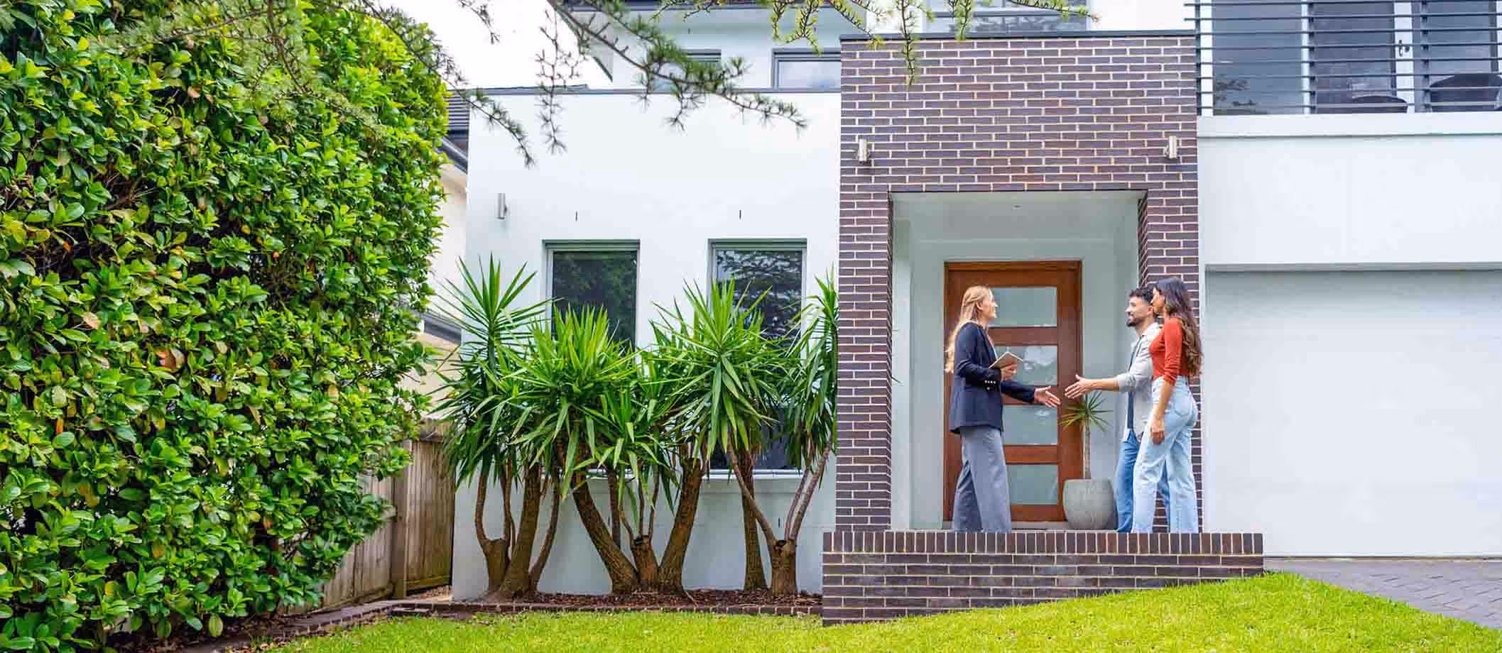 Real estate agent greeting a couple at the entrance of a modern house with brick and white exterior.