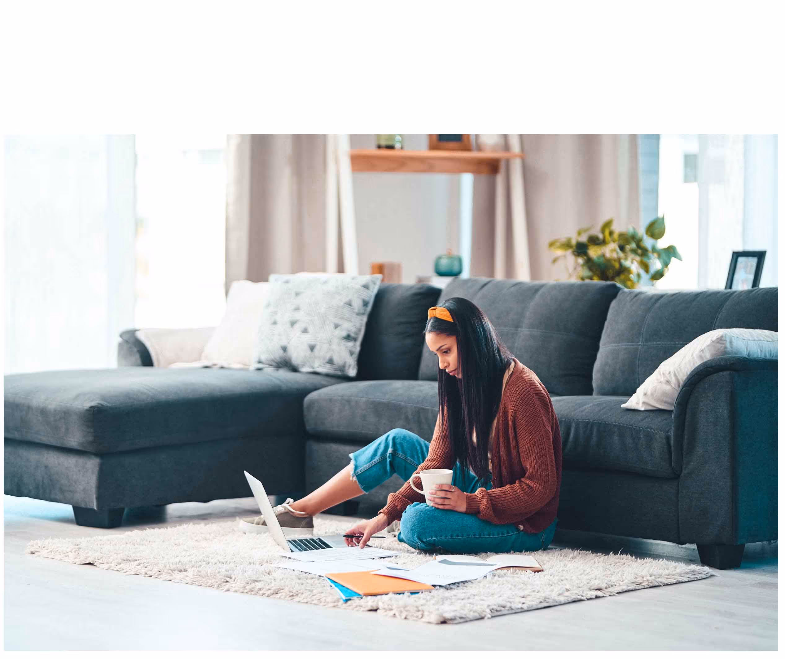 Young woman sitting cross-legged on a rug in living room, using a laptop and holding a mug with documents spread out.