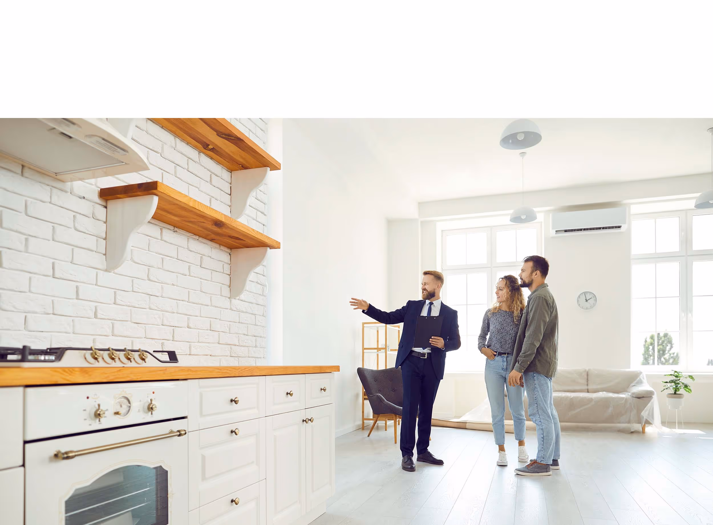 Real estate agent showing a bright, modern kitchen with white cabinets and wooden shelves to a couple in a spacious living area.