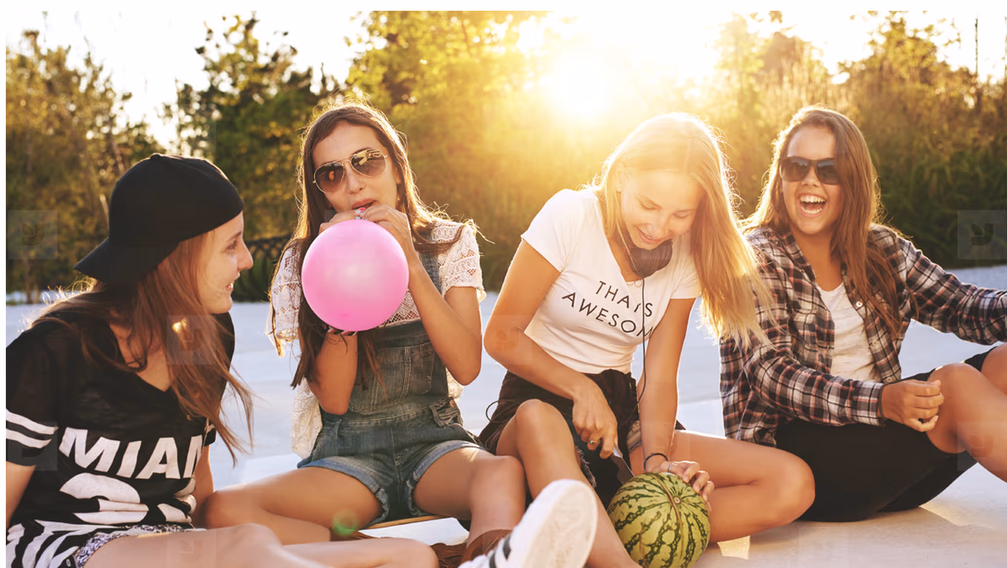 Four teenage girls sitting outdoors in sunlight, one blowing a pink balloon and another cutting a watermelon, all appearing happy.