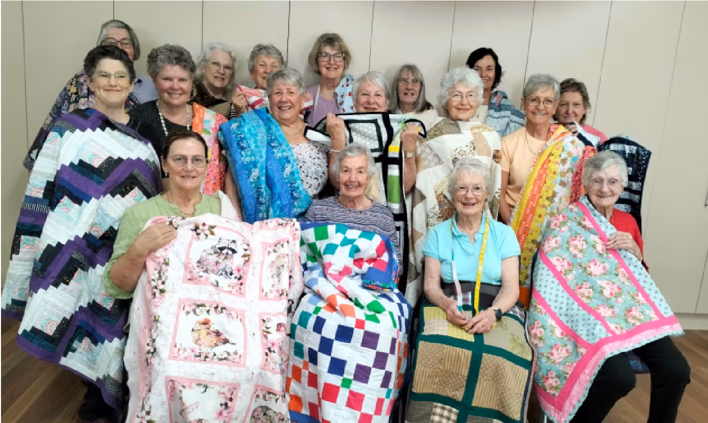 Group of older women smiling and displaying colorful handmade quilts indoors.