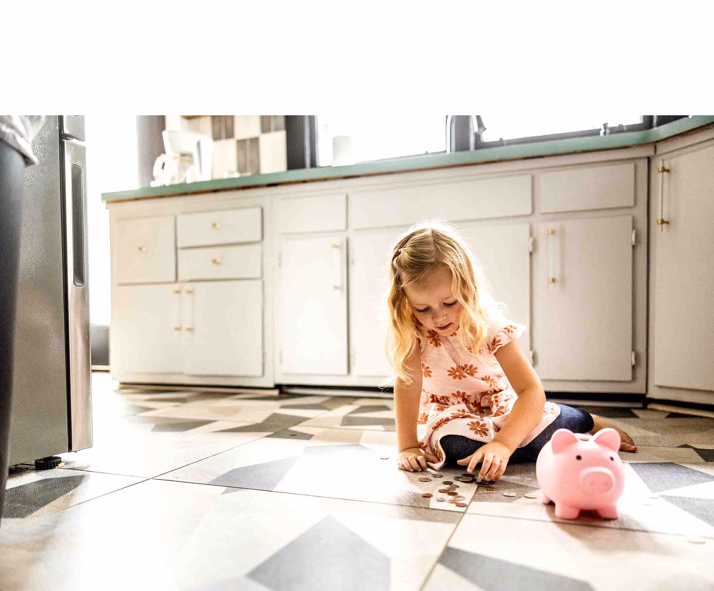 Young girl sitting on kitchen floor counting coins with pink piggy bank nearby.
