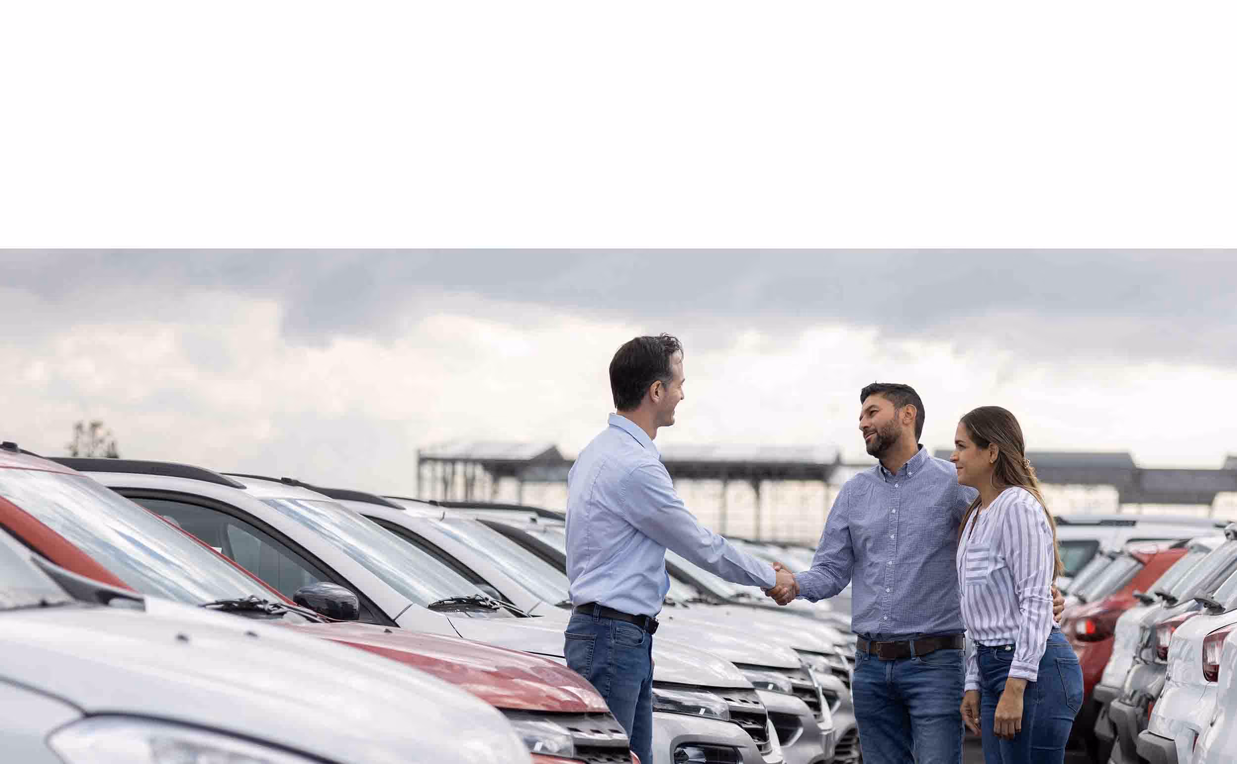 A man shaking hands with a car dealer while a woman stands beside him among rows of parked cars under a cloudy sky.