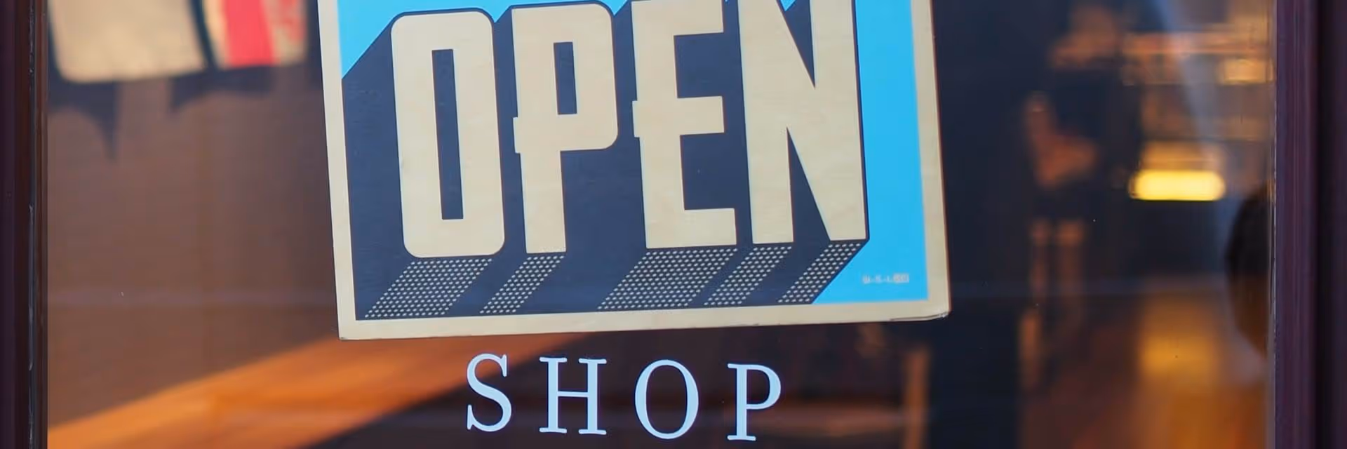 Blue and beige sign reading OPEN hanging above the word SHOP on a glass door.