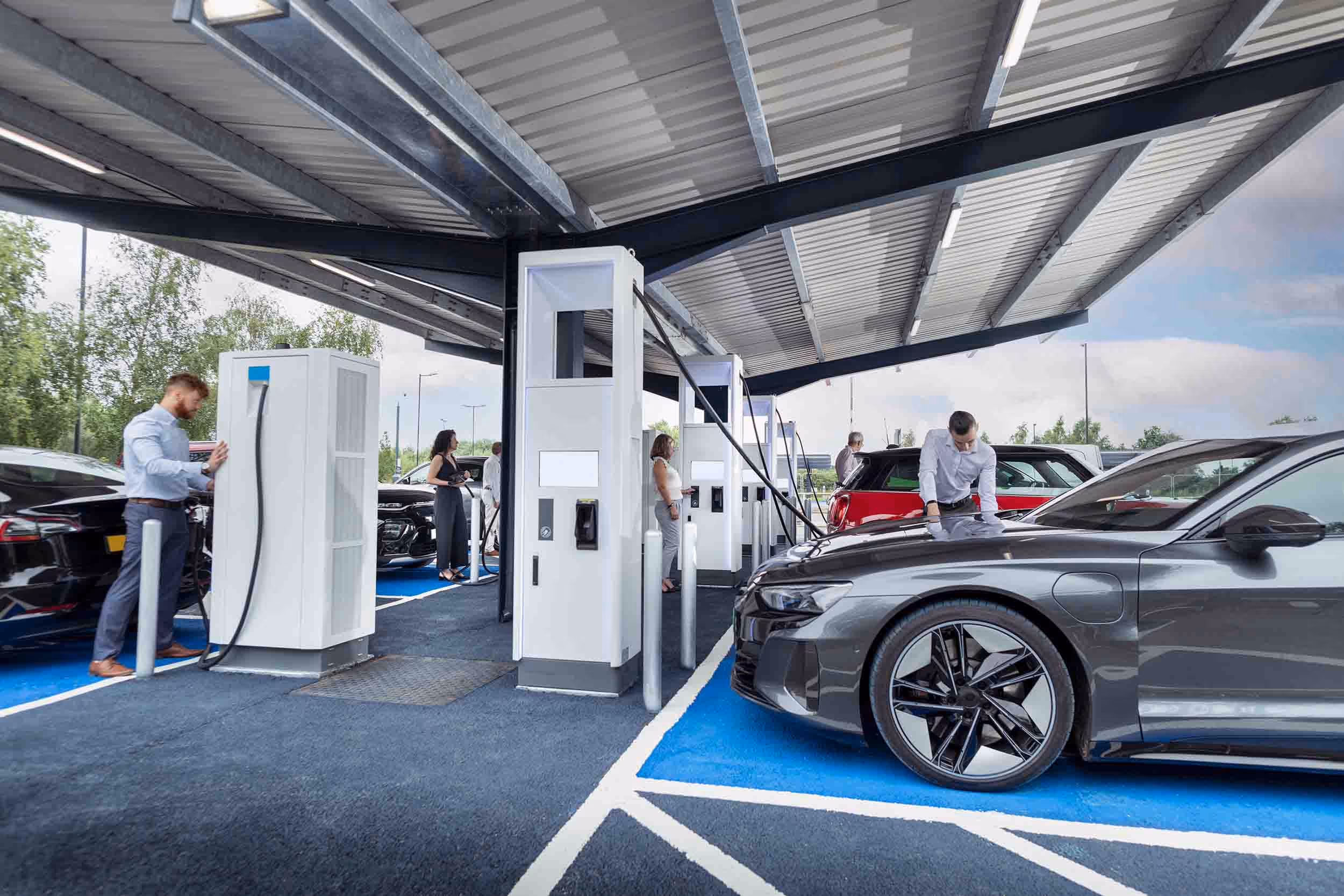 Multiple electric cars charging at a covered station with people attending to them.