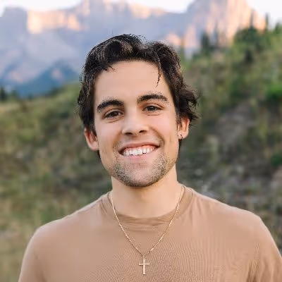 Young man with dark hair wearing a tan shirt and a gold cross necklace smiling outdoors with blurry mountains and greenery in the background.